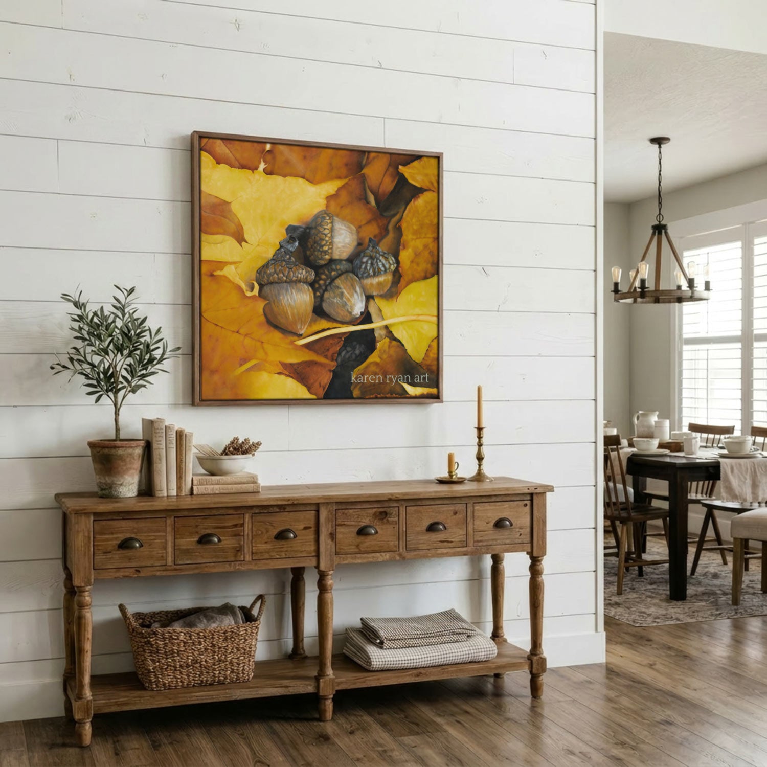 A framed art print depicting acorns on warm yellow and orange autumn leaves, displayed on a white shiplap wall above a rustic wooden console table in a dining room setting.