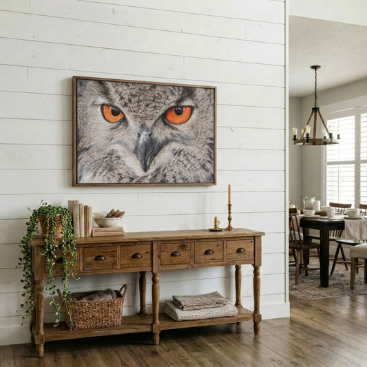 A framed wall art print of an owl with bright orange eyes hanging above a wooden console table in a bright, modern dining room with white shiplap walls.