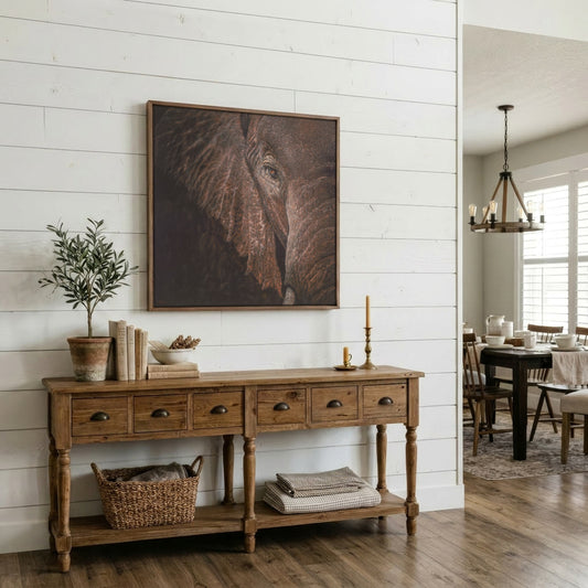 A large framed square art print depicting a close-up of an elephant's face, styled in a rustic living room setting above a wooden console table.