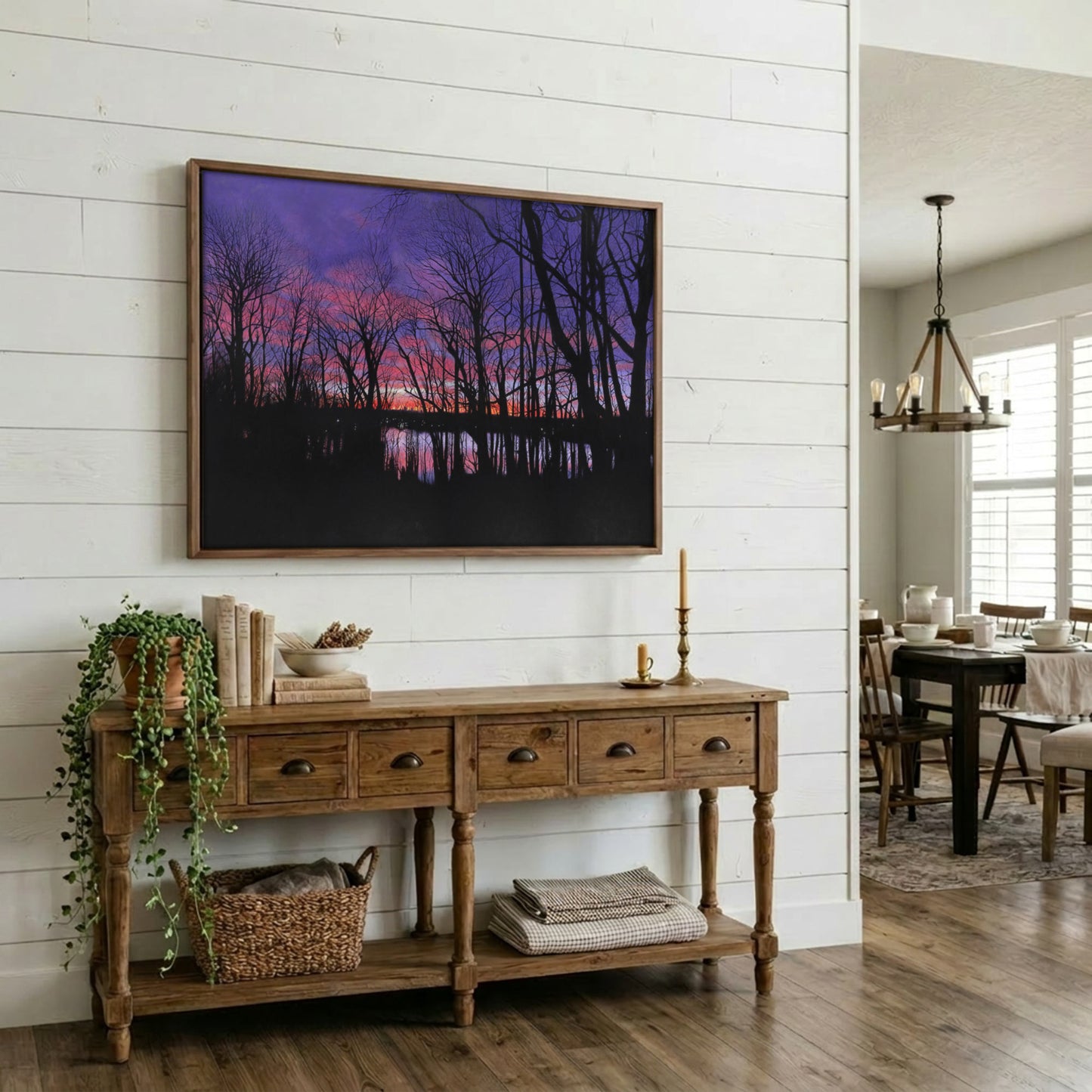 A framed landscape art print depicting a purple and pink sunset over a dark forest and water, displayed on a white shiplap wall above a rustic wooden console table in a styled home interior.