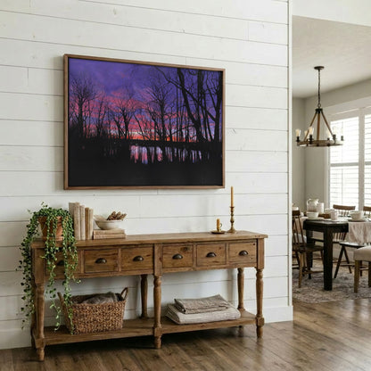 A framed landscape art print depicting a purple and pink sunset over a dark forest and water, displayed on a white shiplap wall above a rustic wooden console table in a styled home interior.