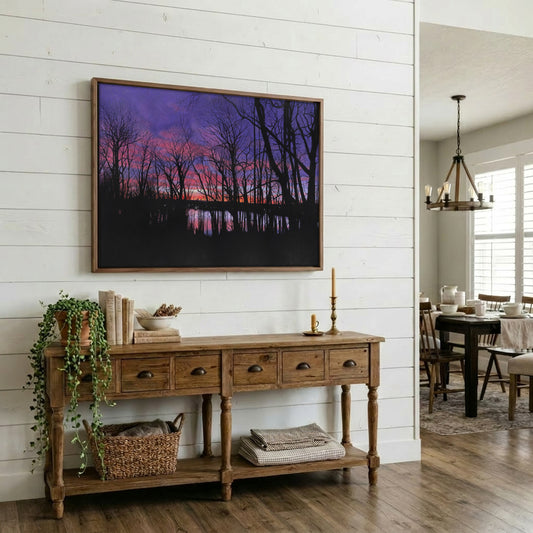 A framed landscape art print depicting a purple and pink sunset over a dark forest and water, displayed on a white shiplap wall above a rustic wooden console table in a styled home interior.